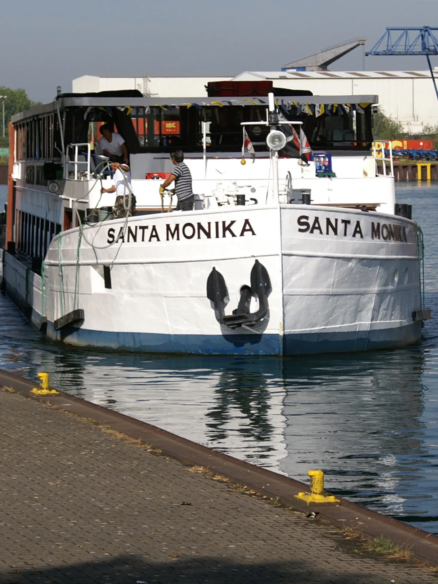 Das weiße Passagierschiff "Santa Monika" liegt im Hafen und ist an der Anlegestelle vertäut. Auf dem Deck sind Besatzungsmitglieder bei der Arbeit. Der Schiffsrumpf reflektiert sich im ruhigen Wasser des Hafens, während im Hintergrund Hafengebäude zu sehen sind.