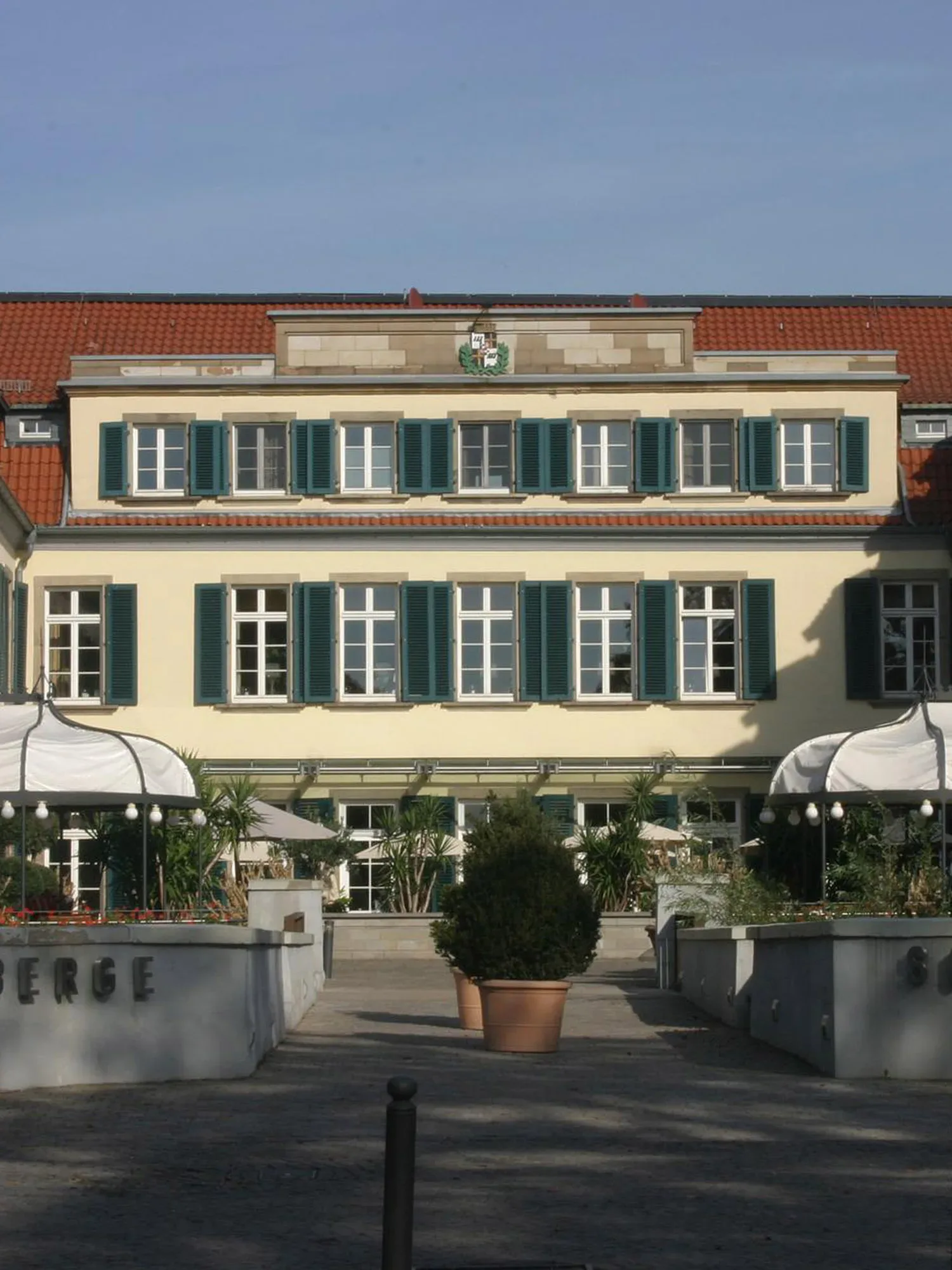 Ein historisches Gebäude mit cremefarbener Fassade und grünen Fensterläden. Vor dem Haupteingang befindet sich eine gepflegte Gartenanlage mit zwei großen Topfpflanzen und überdachten Sitzbereichen. Ein Wappen ziert die Mitte des Dachs.
