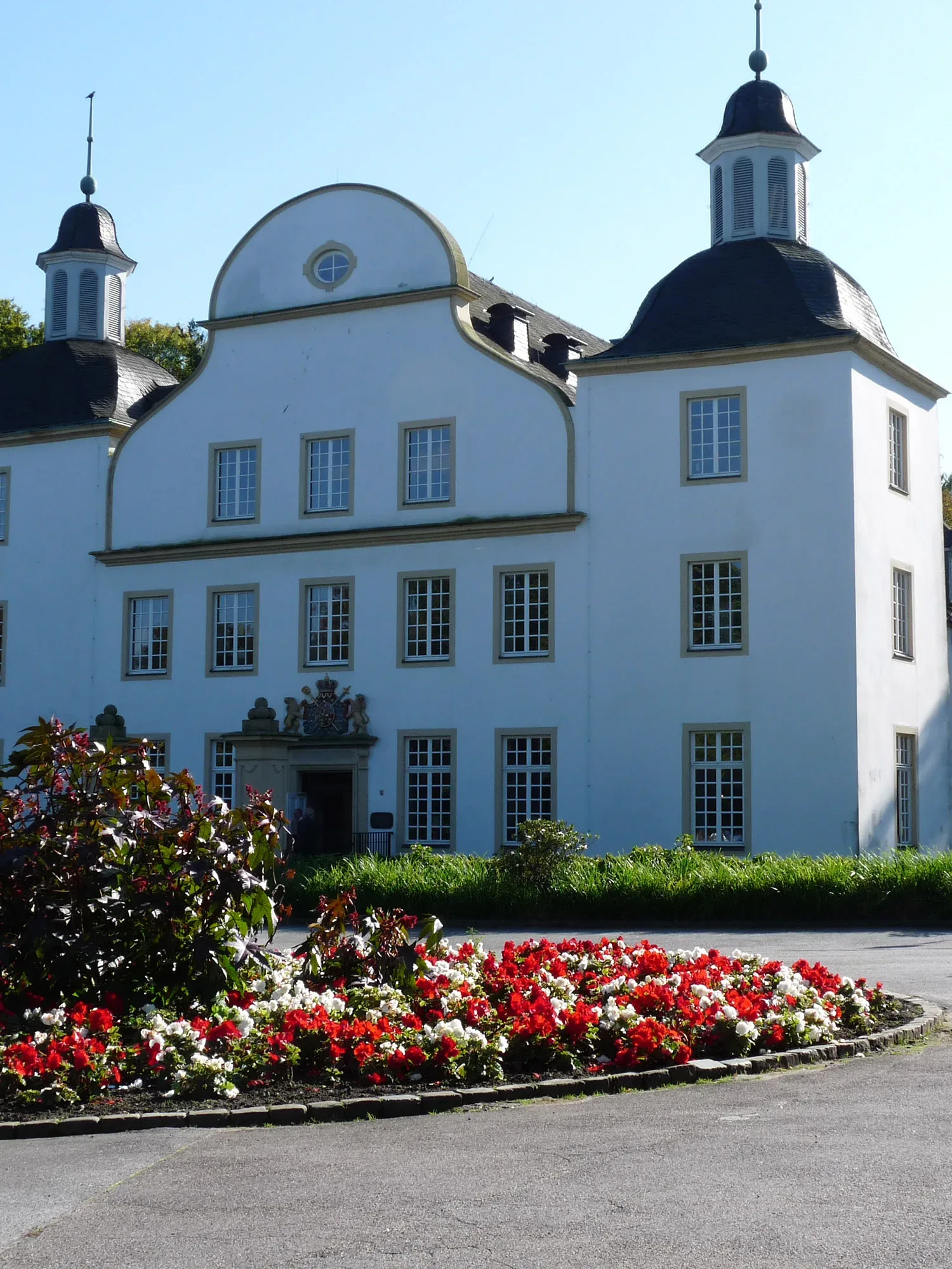 Elegantes weißes Schloss mit zwei symmetrischen Türmen und einem geschwungenen Mittelgiebel. Im Vordergrund blüht ein großes, buntes Blumenbeet, das die schlichte, helle Fassade des Gebäudes ergänzt und einen einladenden Eindruck hinterlässt.
