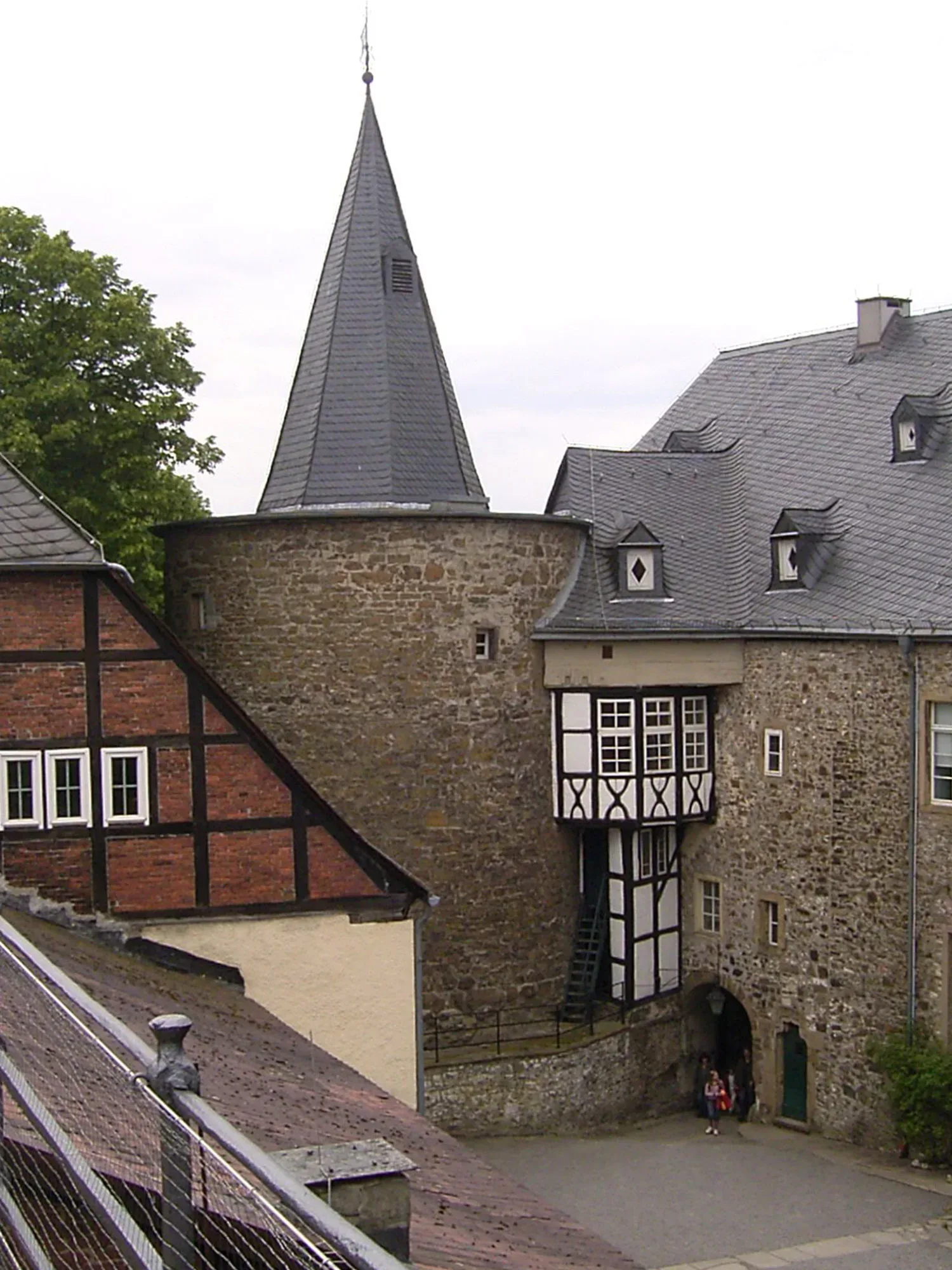 Eine mittelalterliche Burg mit rundem Steinturm und spitzem Schieferdach. Ein Fachwerkhaus schließt sich an den Turm an, und ein Torbogen führt in den Innenhof, in dem zwei Besucher zu sehen sind. Die Burgmauern bestehen aus grobem Stein und erzeugen eine rustikale Atmosphäre.