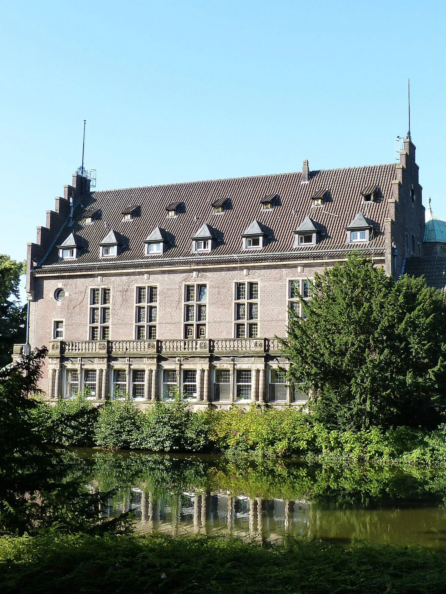 Ein elegantes Schloss mit hohen Fenstern und einem grauen Walmdach, das sich über einem klaren Wassergraben erhebt. Die Fassade spiegelt sich im ruhigen Wasser, und dichte grüne Büsche und Bäume umgeben das Gebäude. Der blaue Himmel verstärkt die friedliche Szene.