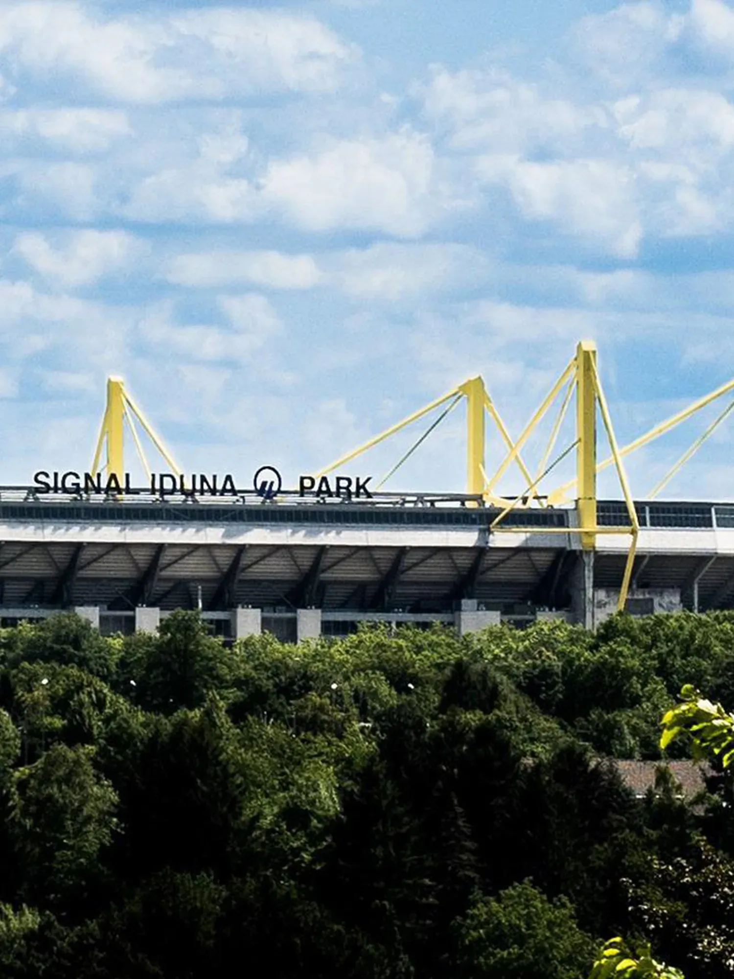 Signal Iduna Park, das Stadion von Borussia Dortmund, ragt über einem dichten Wald auf. Die markanten gelben Pylonen des Stadions heben sich deutlich vor dem blauen Himmel mit weißen Wolken ab. Der Stadionname ist auf der oberen Fassade gut sichtbar.