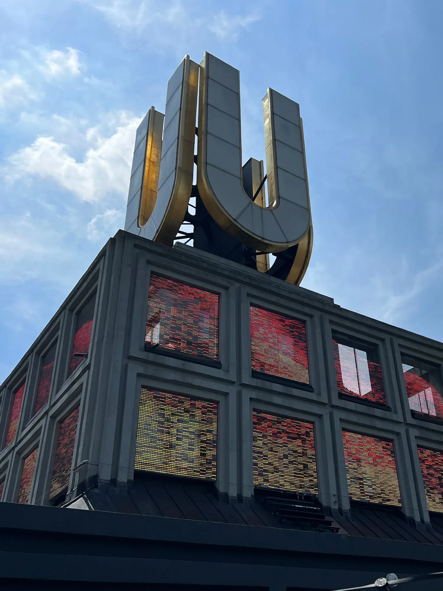 Modernes Gebäude mit grossem, goldfarbenem "U"-Kunstzeichen auf dem Dach. Die Fassade besteht aus Glasflächen, die bunte Lichter reflektieren. Der Himmel im Hintergrund ist blau und klar, was das glänzende "U" hervorhebt.