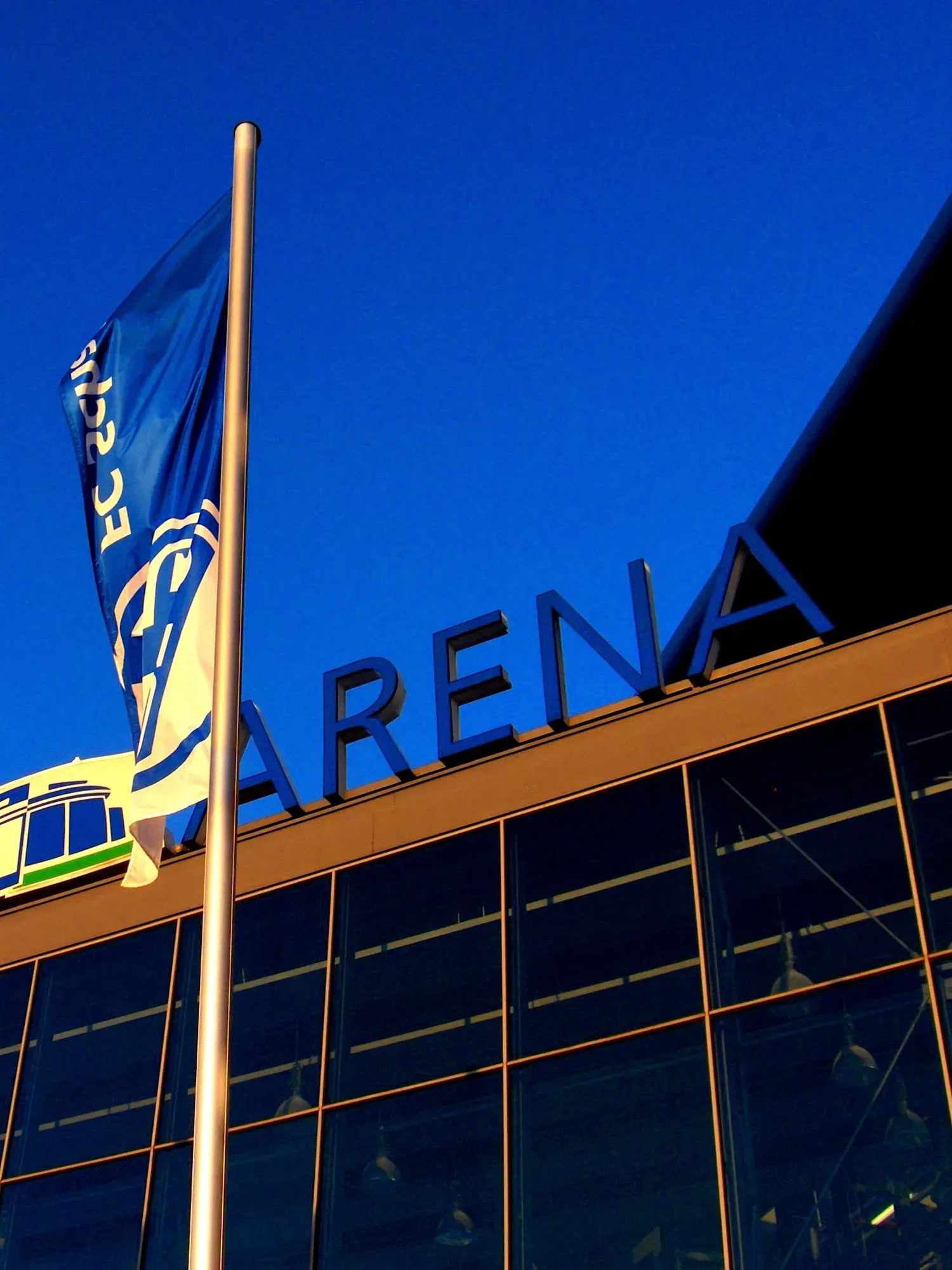 Moderne Arena mit großer Glasfassade, darüber der Schriftzug „Arena“ in minimalistischer Schrift. Vor der Fassade weht eine blau-weiße Flagge mit dem Logo des Fußballvereins Schalke 04. Der Himmel ist tiefblau und strahlt Lebendigkeit aus.
