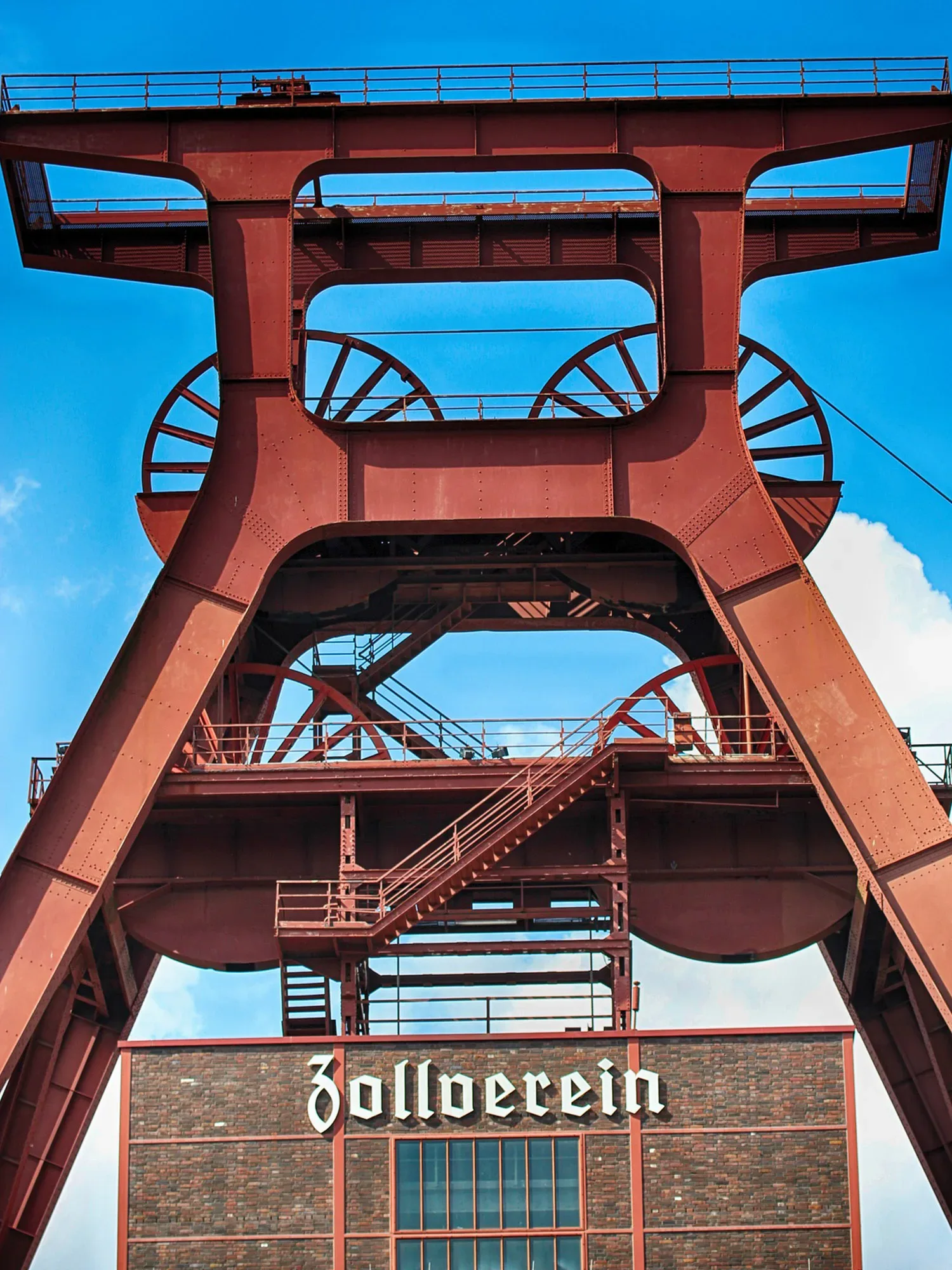 Industrieller Förderturm der Zeche Zollverein in dunklem Rot, mit zwei großen Seilscheiben und robusten Stahlträgern. Die moderne Inschrift „Zollverein“ prangt auf dem Gebäude darunter. Der strahlend blaue Himmel verleiht dem Bauwerk eine markante Silhouette.