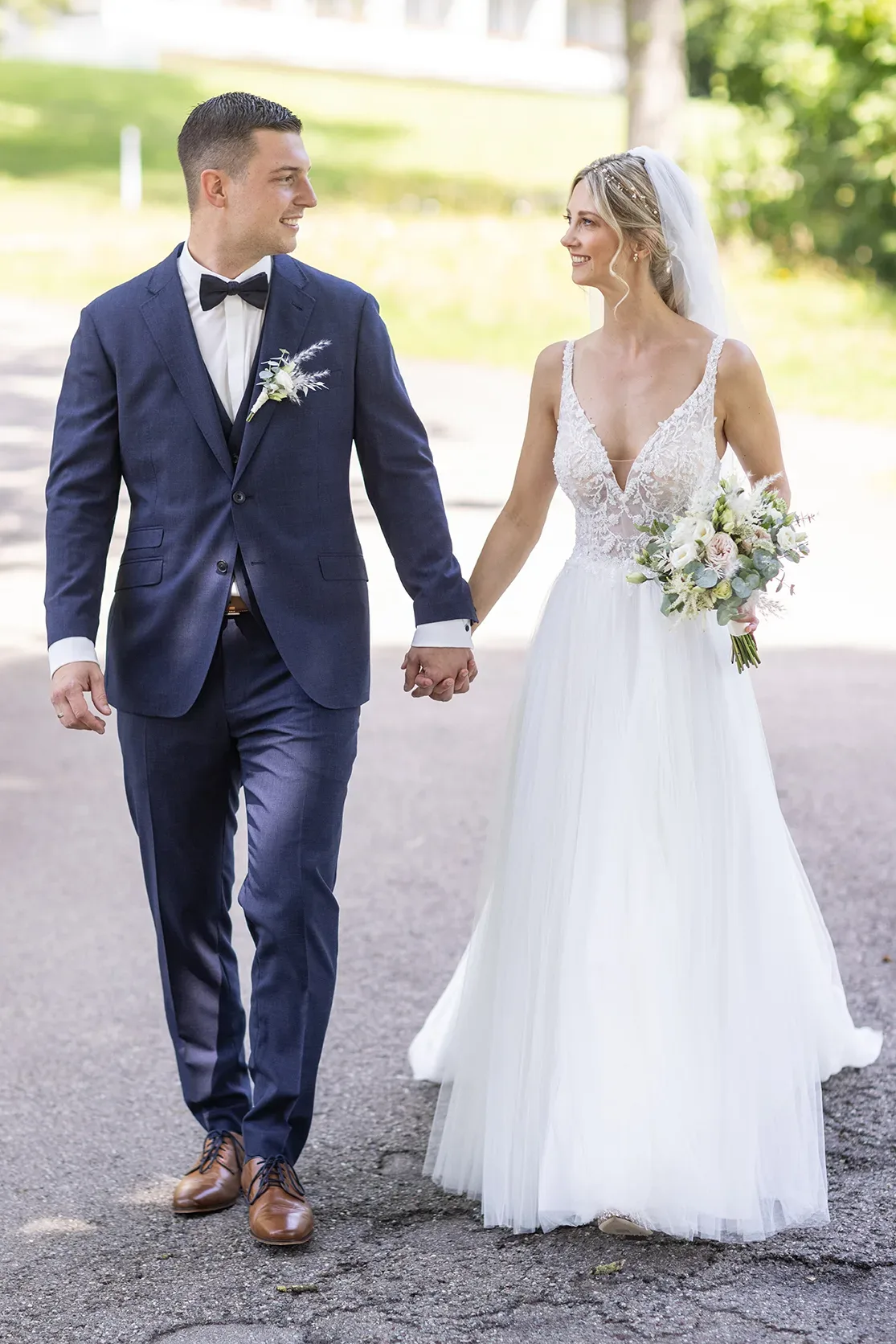 Braut und Bräutigam, Hand in Hand, spazieren an ihrem Hochzeitstag. Die Braut trägt ein Spitzenkleid mit tiefem Ausschnitt, der Bräutigam einen eleganten blauen Anzug.