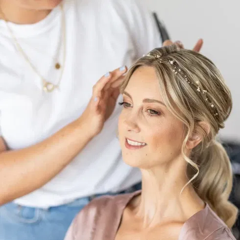 Die Braut bekommt von der Stylistin die letzten Details an ihrer Hochzeitsfrisur mit einem zarten Haarband aus Perlen im Getting-ready-Moment.