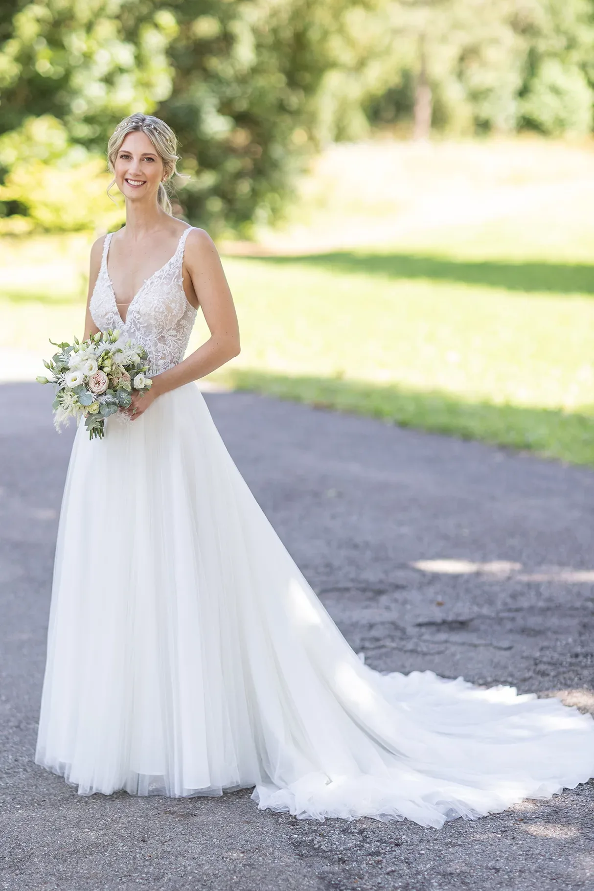 Braut in einem eleganten, bodenlangen Spitzenkleid mit Schleppe, steht lächelnd auf einer sonnigen Straße und hält einen Blumenstrauß.