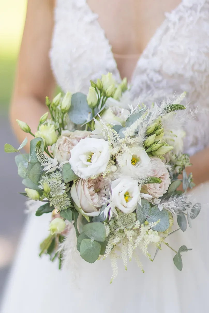 Nahaufnahme eines Brautstraußes mit weißen Rosen, Eukalyptus und zarten grünen Akzenten, den die Braut in einem Spitzenkleid hält.