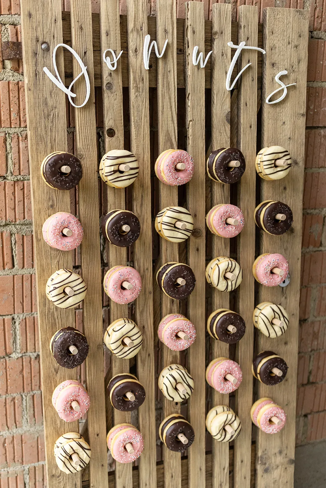 Eine hölzerne Donut-Wand mit verschieden verzierten Donuts in Schokolade, Vanille und rosa Glasur, arrangiert für eine Hochzeit.