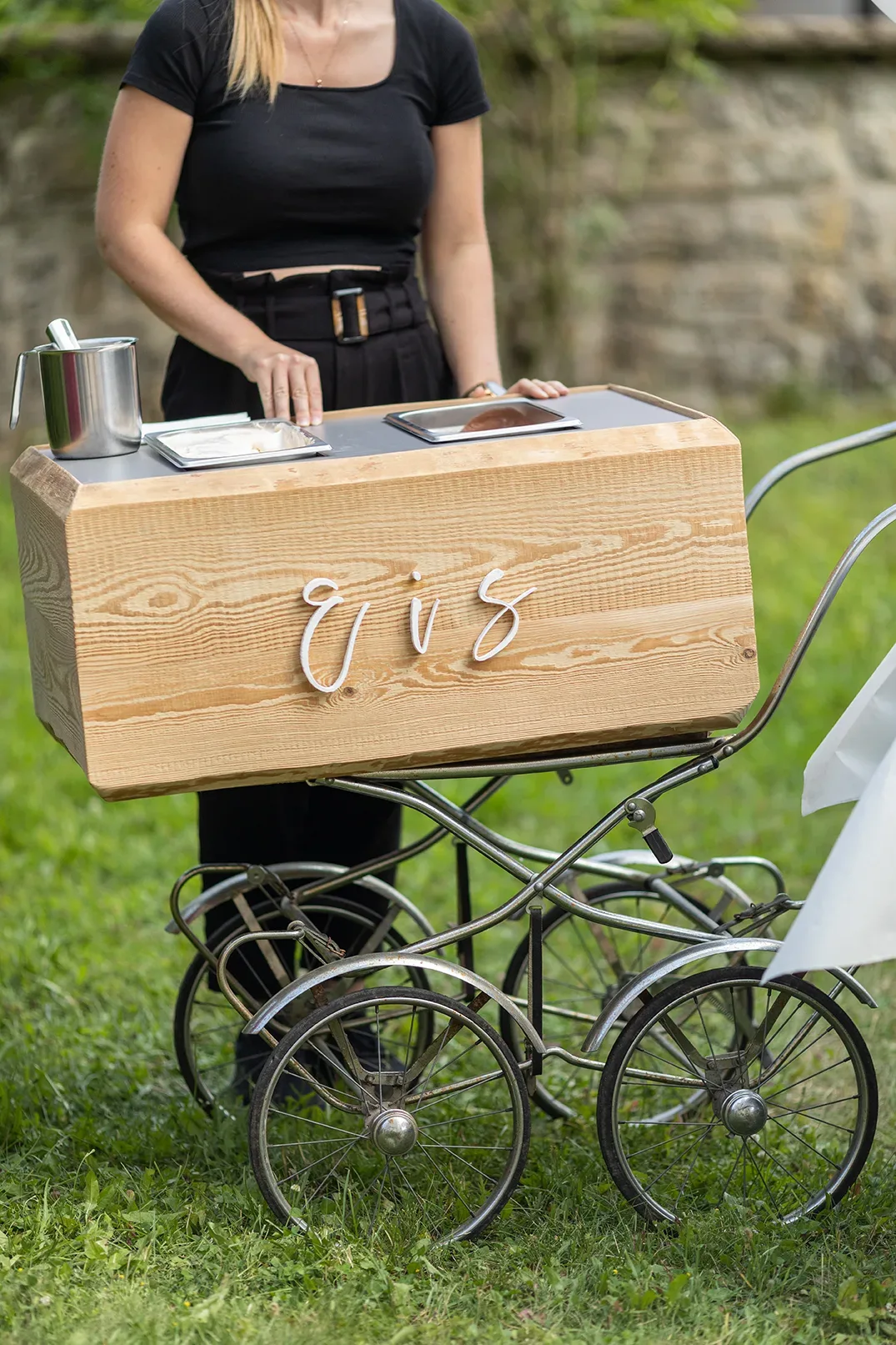 Ein mobiler Eiswagen im Vintage-Stil mit einem handgefertigten Holzaufbau, an dem eine Bedienung Eis für die Hochzeitsgäste serviert.