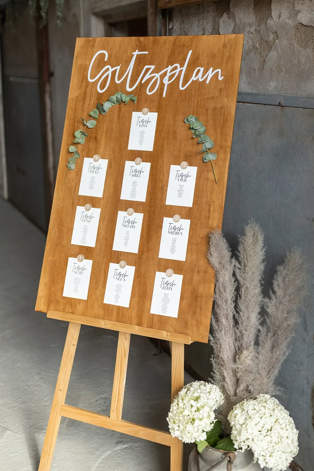 Sitzplan der Hochzeit auf einer Holztafel mit Eukalyptusblättern verziert. Die Tischnummern sind auf weißen Karten gedruckt.