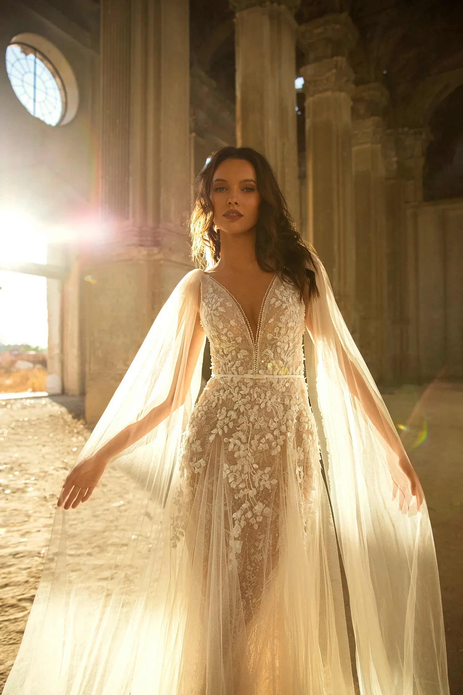 Eine Braut in einem eleganten Hochzeitskleid mit tiefem V-Ausschnitt und langen, fließenden, transparenten Ärmeln posiert in einem von Sonnenlicht durchfluteten Raum.