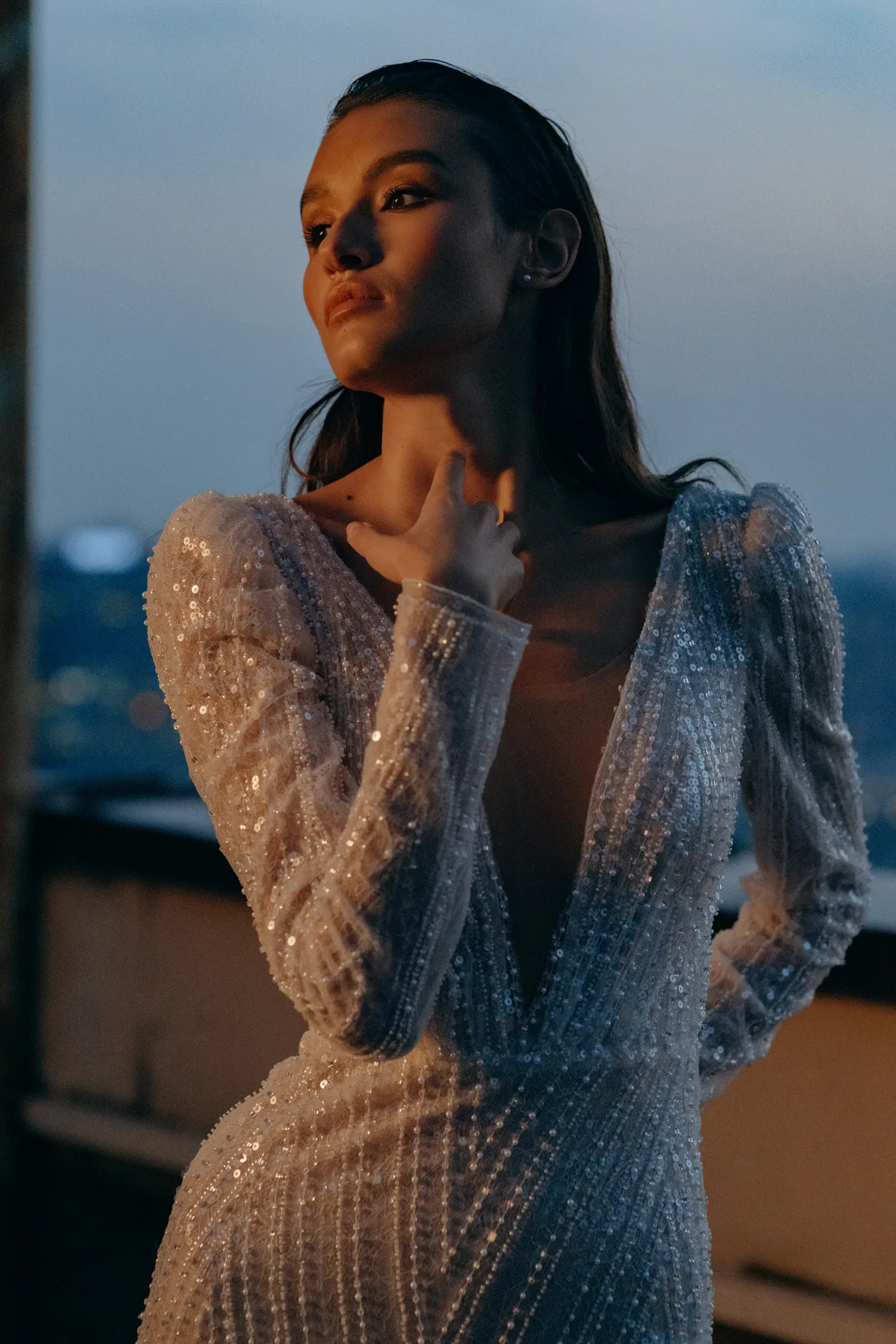 Eine Braut in einem funkelnden Brautkleid mit langen Ärmeln, verziert mit Pailletten und einem tiefen V-Ausschnitt, posiert in einer abendlichen Szenerie.