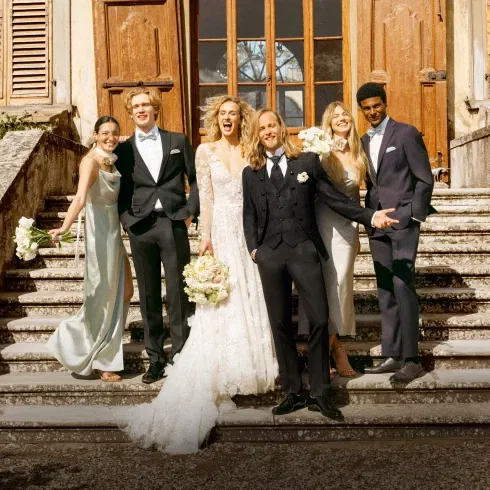 Eine Gruppe festlich gekleideter Hochzeitsgäste und das Brautpaar stehen auf einer eleganten Steintreppe vor einer historischen Location, lächelnd in die Kamera.