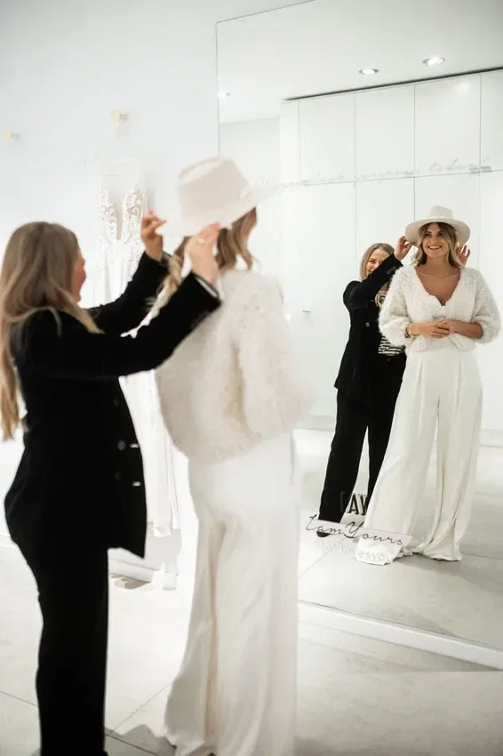 Eine Braut probiert vor einem Spiegel einen weißen Jumpsuit und einen passenden Hut an, während eine Stylistin das Accessoire ausrichtet. Der helle, minimalistische Raum und die Brautmode im Hintergrund schaffen eine stilvolle Atmosphäre.