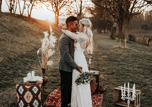 Ein Hochzeitspaar steht auf einer Wiese bei Sonnenuntergang. Die Braut trägt ein langes, weisses Kleid aus Spitze und hält einen kleinen Blumenstrauss, während der Bräutigam in einem grauen Sakko gekleidet ist. Um das Paar herum liegen dekorative Teppiche in Rot- und Erdtönen, und auf einem Holztisch steht eine Vase mit getrocknetem Pampasgras. Die warmen Farben des Sonnenuntergangs beleuchten die Szene und schaffen eine romantische Atmosphäre.