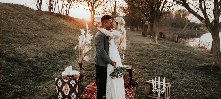 Ein Hochzeitspaar steht auf einer Wiese bei Sonnenuntergang. Die Braut trägt ein langes, weisses Kleid aus Spitze und hält einen kleinen Blumenstrauss, während der Bräutigam in einem grauen Sakko gekleidet ist. Um das Paar herum liegen dekorative Teppiche in Rot- und Erdtönen, und auf einem Holztisch steht eine Vase mit getrocknetem Pampasgras. Die warmen Farben des Sonnenuntergangs beleuchten die Szene und schaffen eine romantische Atmosphäre.