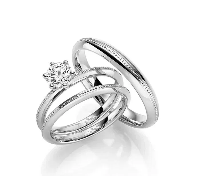 Zwei Weißgold-Eheringe, einer mit einem Solitär-Diamanten im Mittelpunkt und der andere schlicht gehalten. Beide Ringe haben feine, dezente Kanten, die das moderne und minimalistische Design unterstreichen.