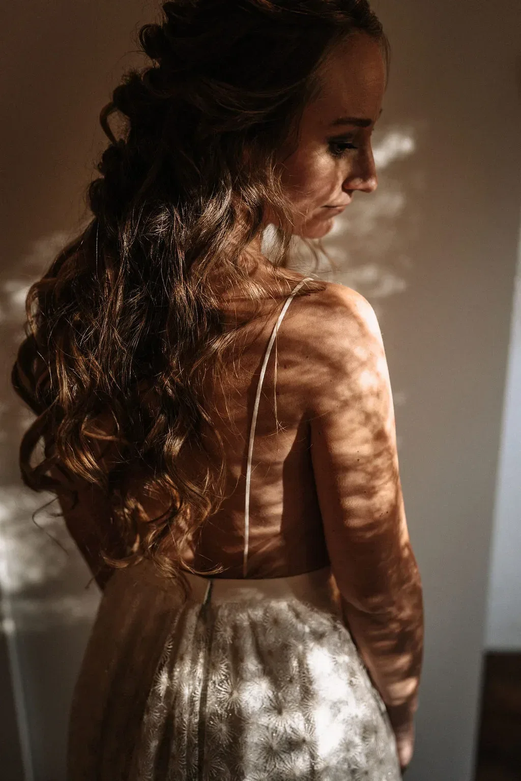 Eine Braut in einem eleganten Kleid mit offenem Rücken, beleuchtet von weichem, schattenhaftem Licht, das ihre Locken und das Spitzenmuster ihres Kleides hervorhebt.