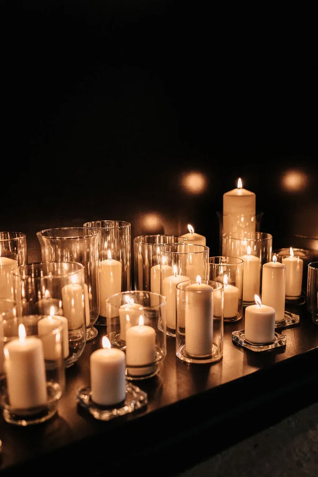Mehrere brennende Kerzen in Glasgefäßen, die in einem stimmungsvollen, dunklen Raum arrangiert sind, erzeugen eine warme und gemütliche Atmosphäre.