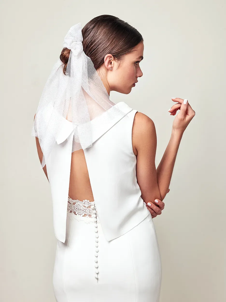 Rückansicht einer Braut in einem eleganten, weißen Kleid mit tiefem Rückenausschnitt und Spitzen-Detail. Ihr Haar ist zu einem Knoten gebunden, verziert mit einem zarten Schleier.