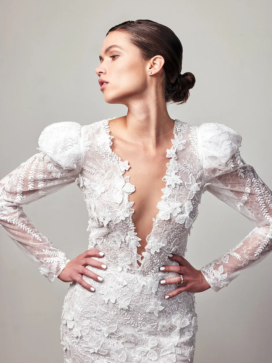 Seitliche Aufnahme einer Braut in einem tief ausgeschnittenen Spitzenkleid mit Puffärmeln. Sie posiert selbstbewusst und schaut zur Seite.