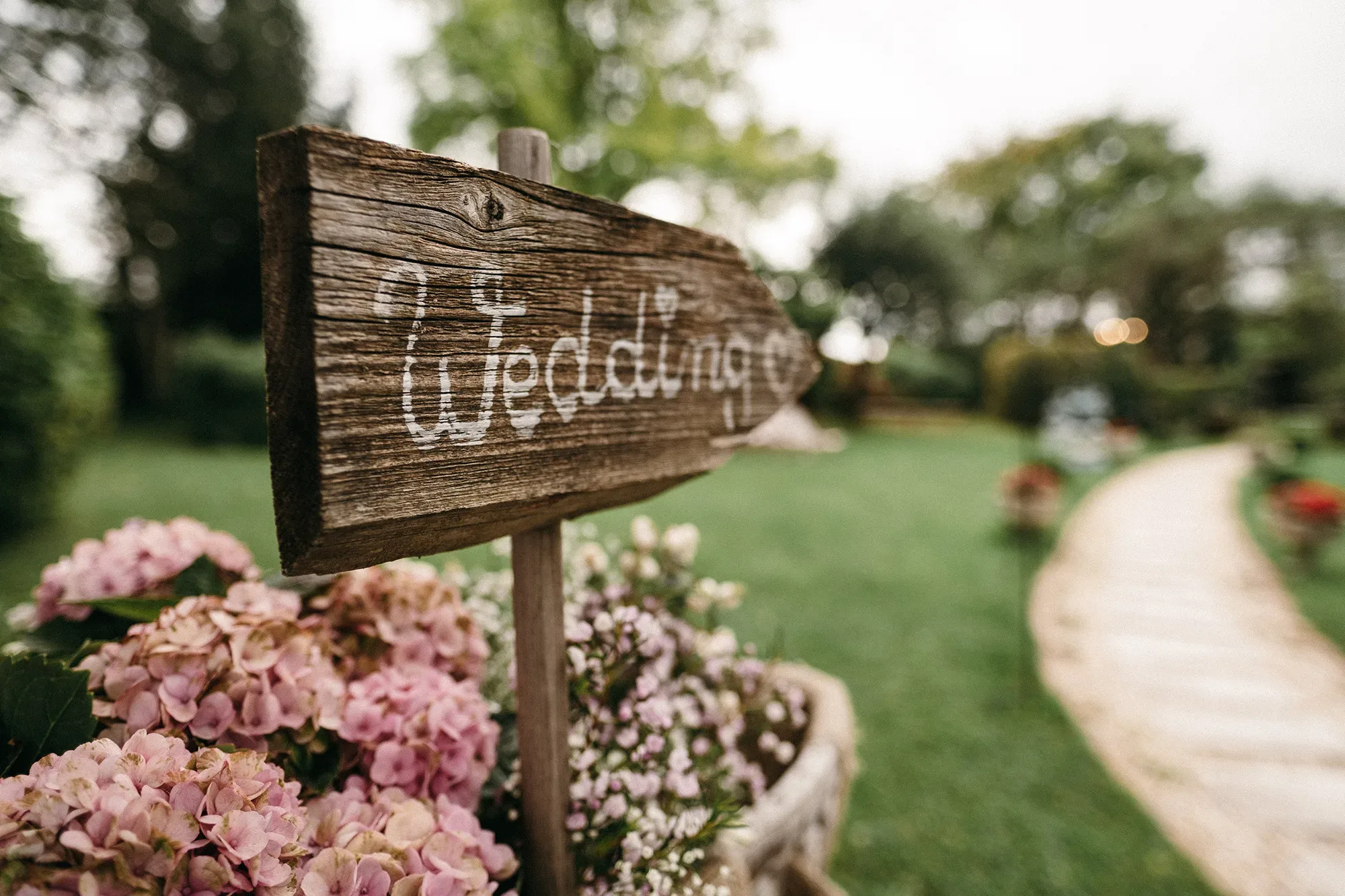 Ein rustikaler Holzwegweiser mit der Aufschrift 'Wedding' steht inmitten eines Gartens, umgeben von blühenden rosa Hortensien und einem gepflasterten Pfad.