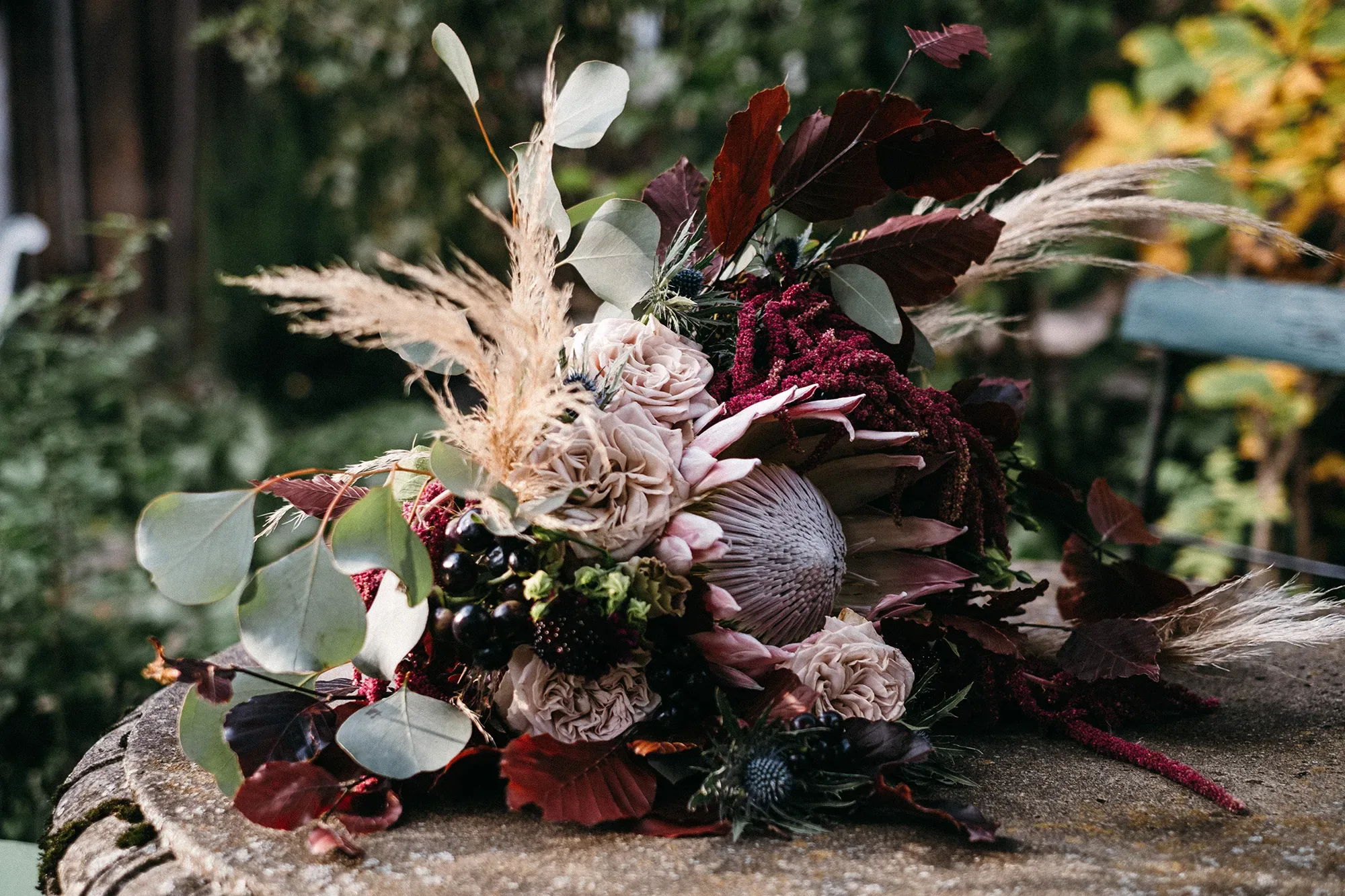Ein üppiges Blumengesteck mit Pampasgras, Protea, Eukalyptusblättern und dunklen Beeren auf einem rustikalen Holztisch in einer natürlichen Umgebung.