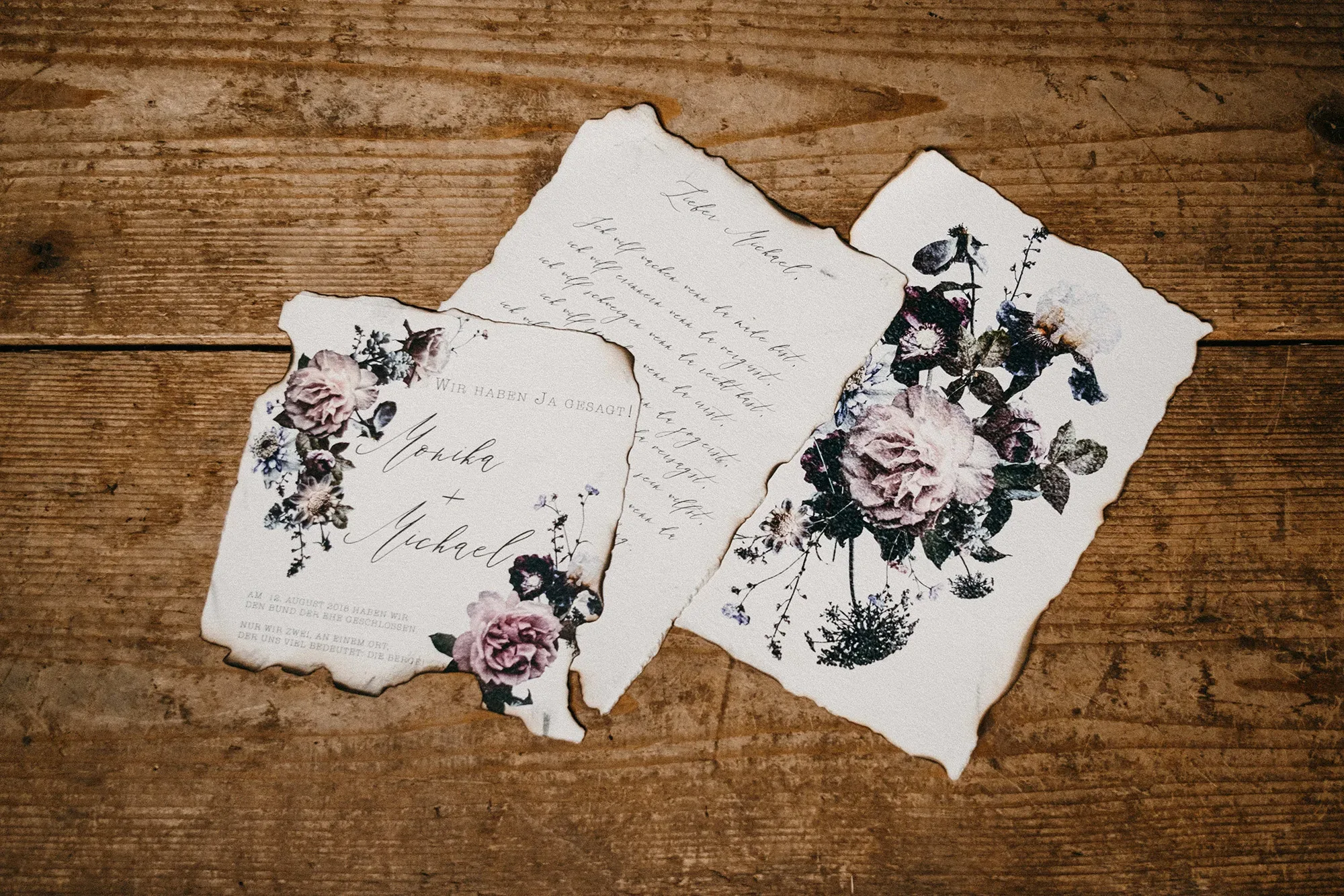 Rustikale Hochzeitseinladungen auf Pergamentpapier mit floralen Motiven und Handlettering, arrangiert auf einem alten Holztisch mit leicht verbrannten Rändern.