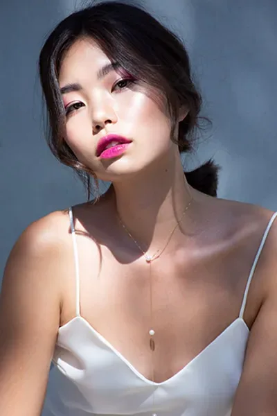Model mit eleganter Hochsteckfrisur und strahlendem Make-up. Sie trägt ein schlichtes, weißes Spaghettiträger-Top, während ihr leuchtender Lippenstift und die feinen Gesichtszüge im Fokus stehen.