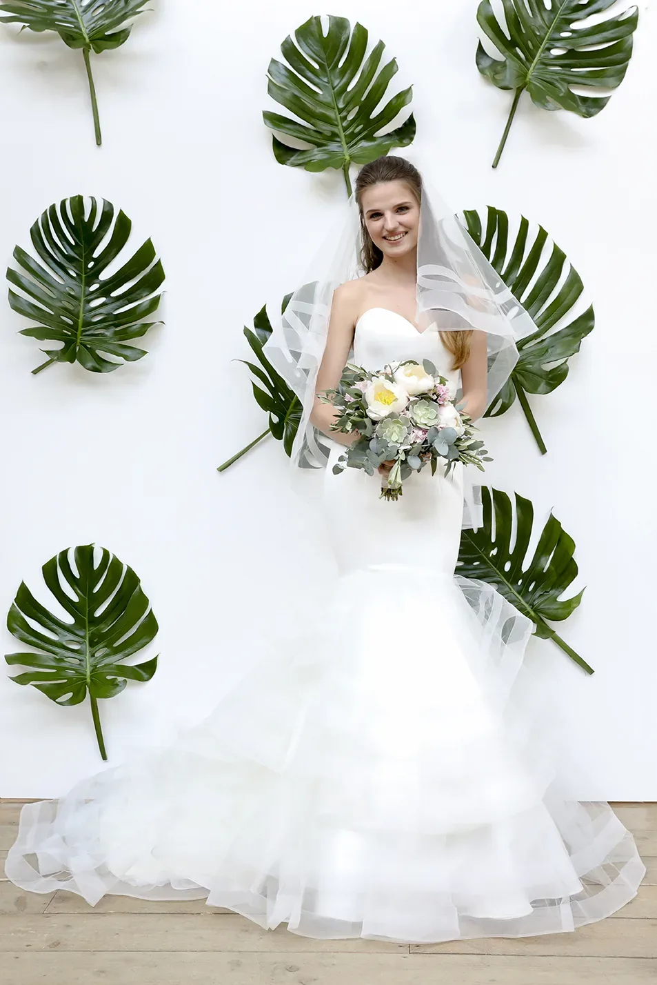 Braut in einem weißen, figurbetonten Kleid mit Rüschenrock und trägerlosem Ausschnitt, steht vor einer weißen Wand, die mit großen Monstera-Blättern dekoriert ist. Sie hält einen üppigen Blumenstrauß und trägt einen zarten Schleier, der locker auf ihre Schultern fällt.