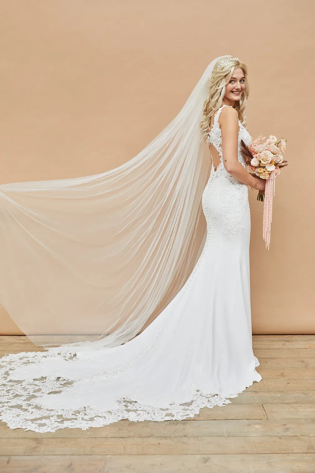 Blonde Braut in einem weißen, eng anliegenden Brautkleid mit floraler Spitze und einem langen Schleier, der dramatisch über den Boden fällt. Sie hält einen pastellfarbenen Blumenstrauß und steht vor einem beigen Hintergrund.