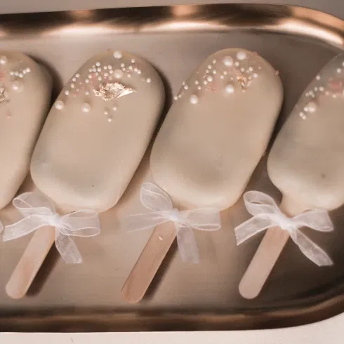 Vier kunstvoll verzierte Cake Popsicles mit weißem Überzug, dekoriert mit essbaren Perlen und Blattgold, liegen auf einem goldenen Tablett. Die Stiele der Popsicles sind mit kleinen, weißen Schleifen geschmückt, die den edlen Look unterstreichen.