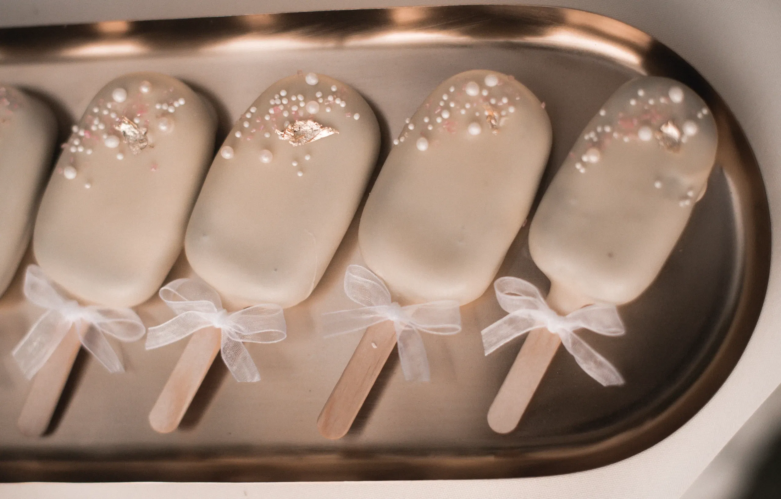 Vier kunstvoll verzierte Cake Popsicles mit weißem Überzug, dekoriert mit essbaren Perlen und Blattgold, liegen auf einem goldenen Tablett. Die Stiele der Popsicles sind mit kleinen, weißen Schleifen geschmückt, die den edlen Look unterstreichen.