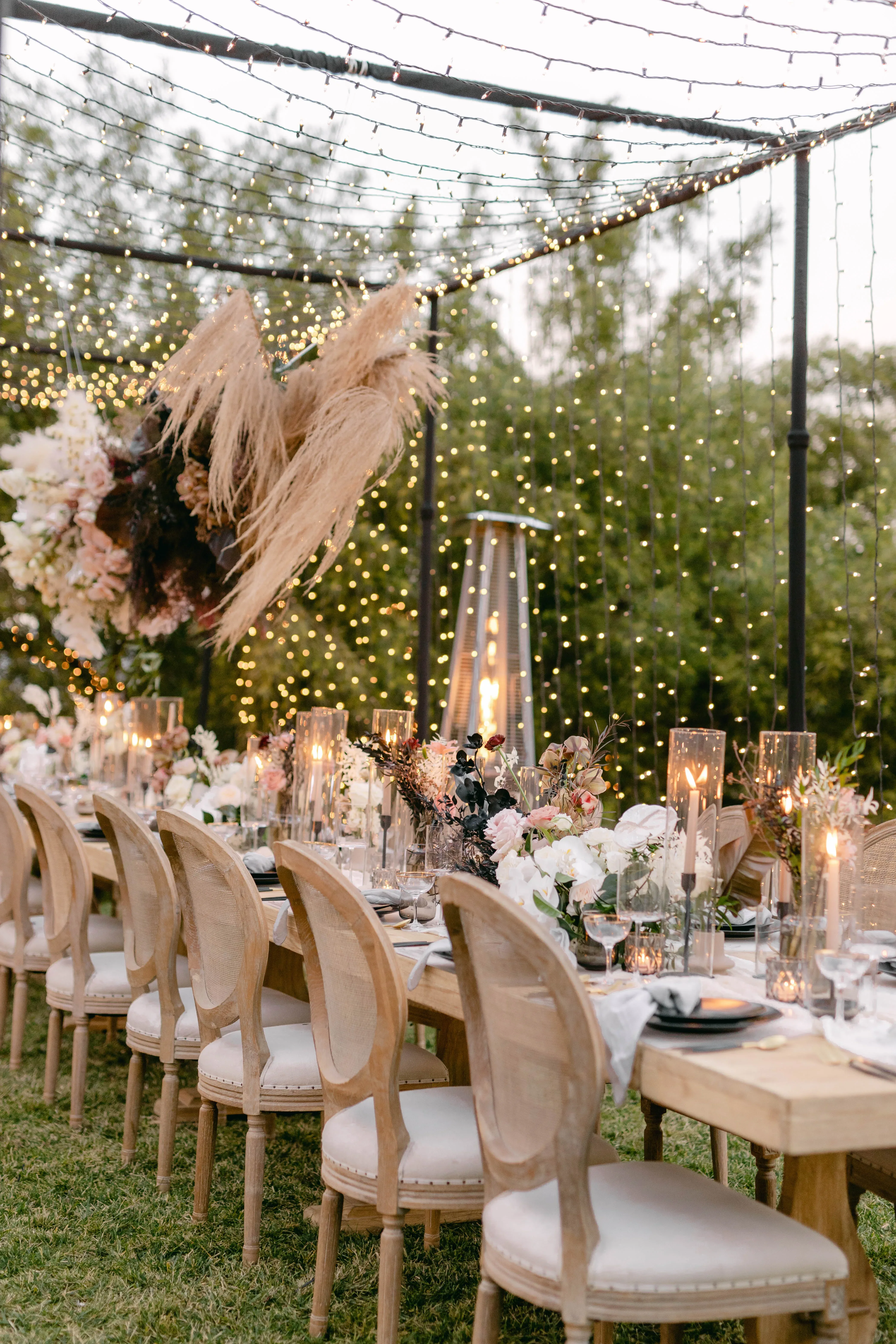Ein langer, festlich gedeckter Tisch bei einer Outdoor-Hochzeit, beleuchtet von Kerzen und umgeben von Lichterketten, sorgt für eine romantische Atmosphäre.