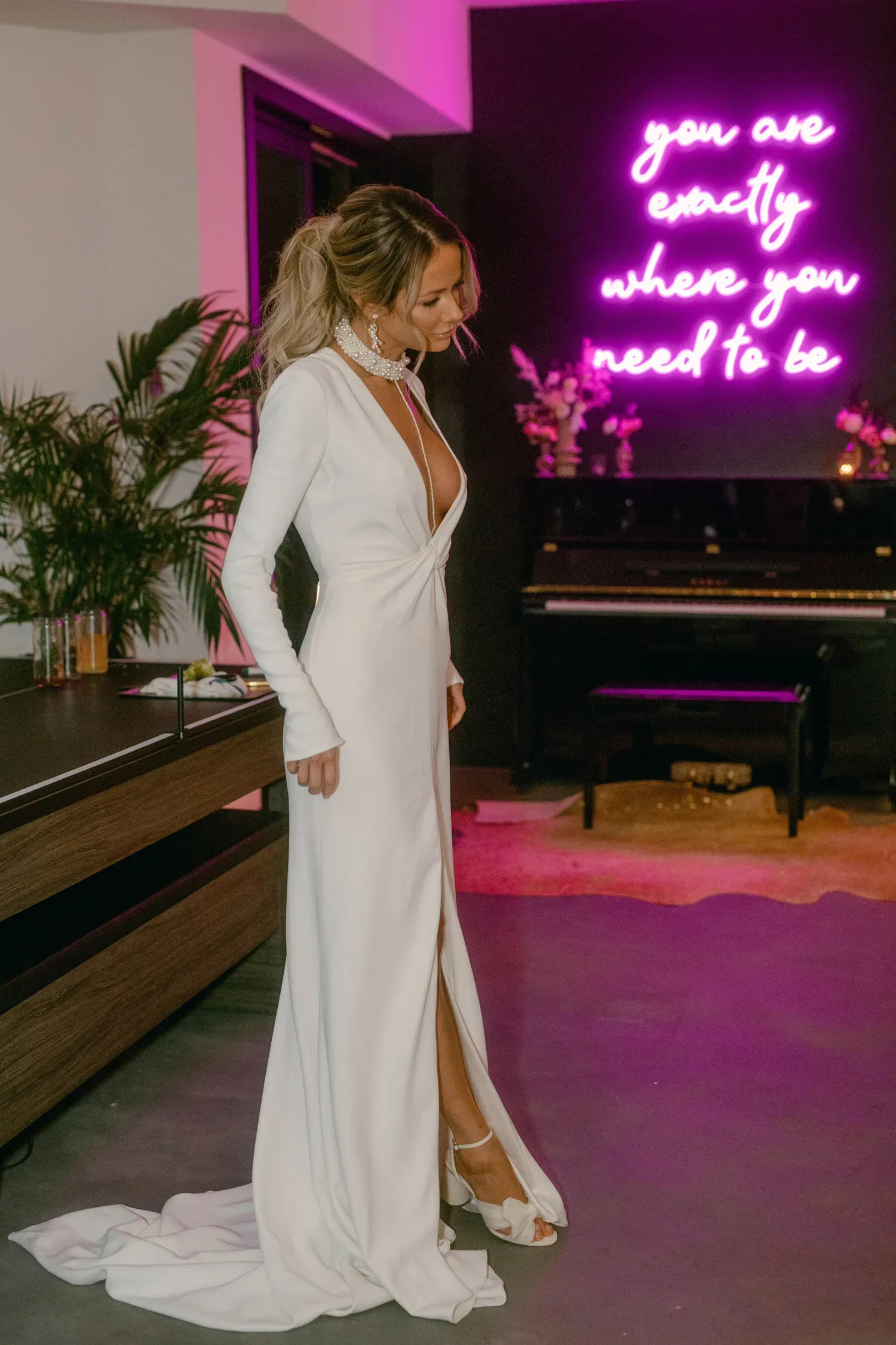 Eine Braut in einem eleganten, tief ausgeschnittenen Hochzeitskleid steht vor einer modernen Neonwand mit der Aufschrift "You are exactly where you need to be".