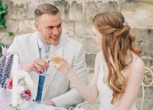 Braut und Bräutigam stoßen bei ihrer Hochzeit im Freien mit Champagner an, beide lächeln sich verliebt an.