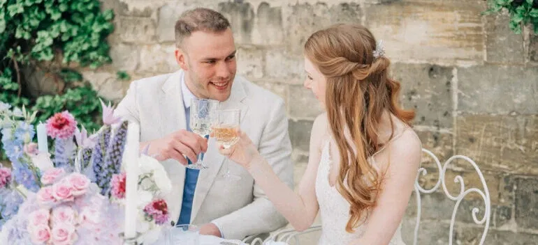 Braut und Bräutigam stoßen bei ihrer Hochzeit im Freien mit Champagner an, beide lächeln sich verliebt an.
