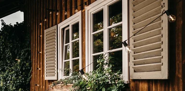 Rustikales Holzhaus mit weißen Fensterläden, Blumenkasten und einer warmen Lichterkette, die eine gemütliche Atmosphäre für eine Hochzeit im Landhausstil schafft.