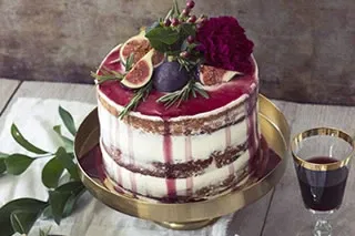 Eine Hochzeitstorte im Naked Cake-Stil mit weißer Glasur und rotem Drip-Effekt, dekoriert mit Feigen, Rosmarin und tiefroten Blumen. Die Torte steht auf einem goldenen Teller.