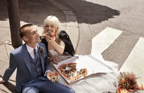 Ein Brautpaar sitzt auf dem Boden und genießt gemeinsam Pizza. Der Bräutigam trägt einen blauen Anzug, während die Braut eine Lederjacke über ihrem Hochzeitskleid trägt.