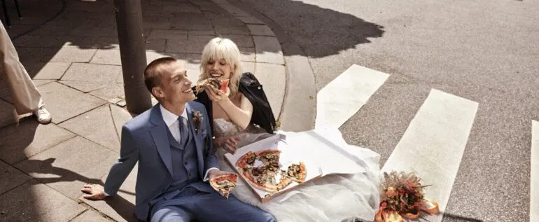 Ein Brautpaar sitzt auf dem Boden und genießt gemeinsam Pizza. Der Bräutigam trägt einen blauen Anzug, während die Braut eine Lederjacke über ihrem Hochzeitskleid trägt.