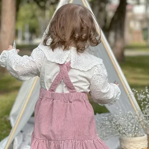 Ein kleines Mädchen von hinten, das eine Spielzeugkrone hält, steht vor einem Tipi-Spielzelt in einem Park. Sie trägt eine weiße Bluse mit Rüschen und einen rosafarbenen Rock.
