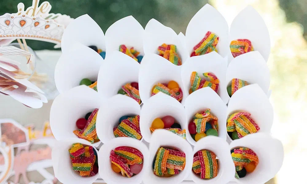Ein Tablett voller kegelförmiger Verpackungen mit bunten Gummibändern und Süßigkeiten, arrangiert auf einem Festtisch mit anderen Süßigkeiten und einer Tiara im Hintergrund.