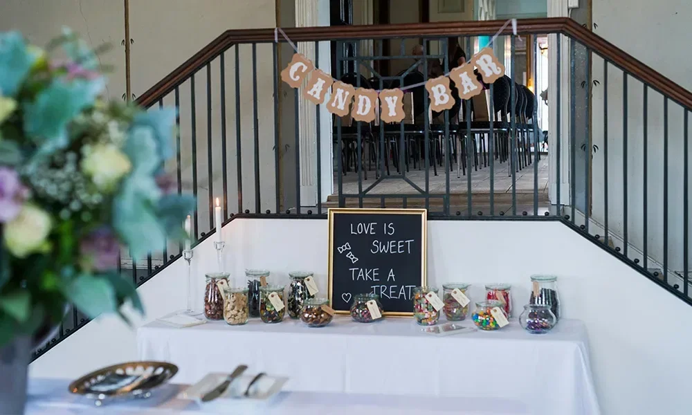 Ein Tisch mit einer Vielfalt von Süßigkeiten in Glasgefäßen steht unter einer Treppe, auf der Tischdecke ein Schild mit der Aufschrift 'Love is sweet, take a treat' neben einem 'Candy Bar'-Banner.