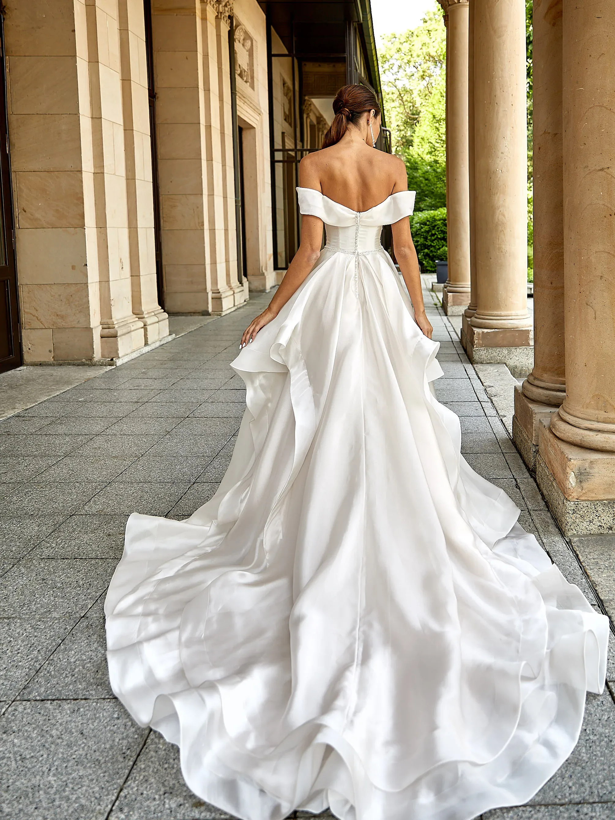 Braut in einem schulterfreien, fließenden Seidenkleid mit langer Schleppe, die durch eine elegante Säulenhalle schreitet.