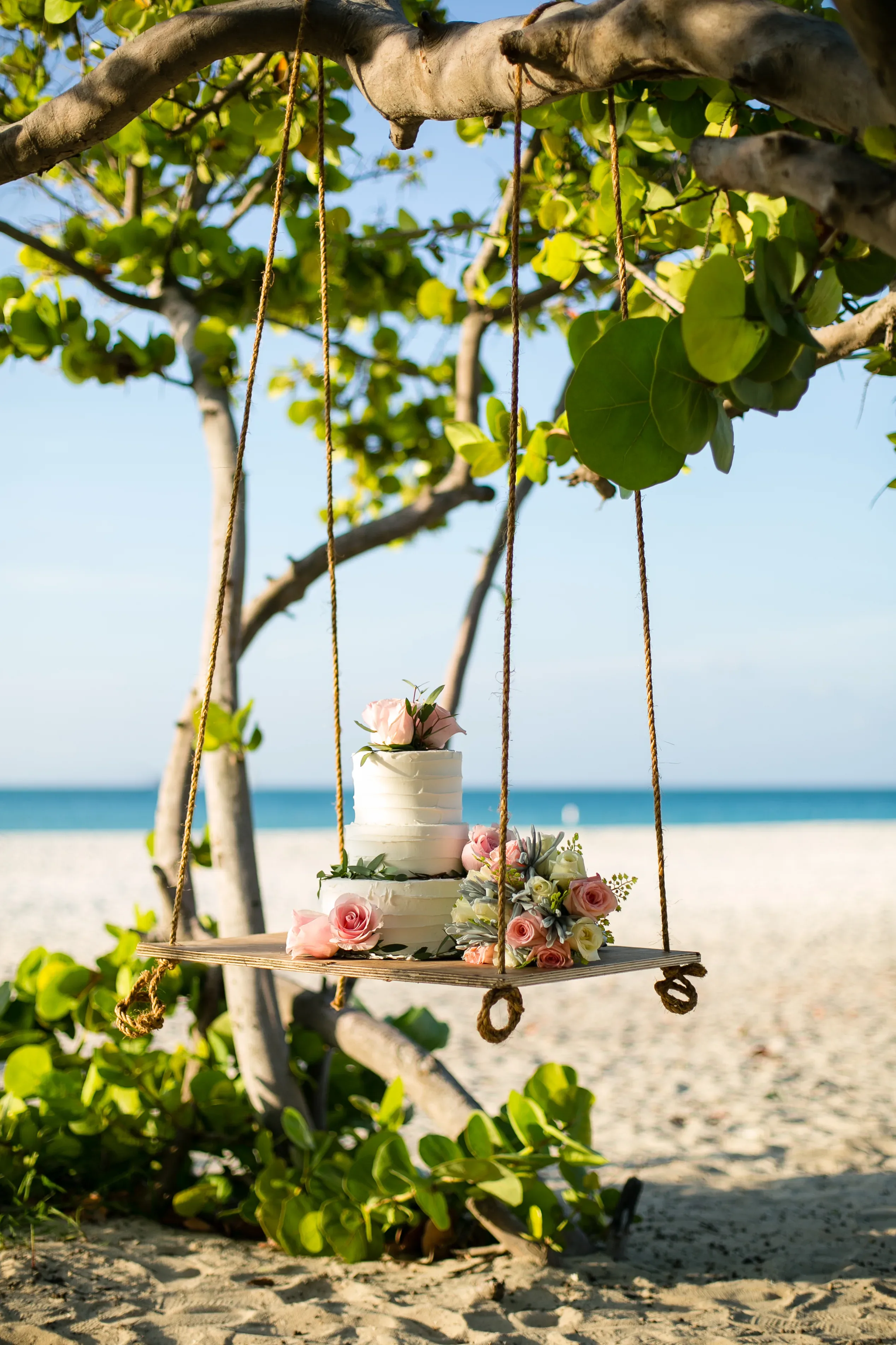 Eine weiße Hochzeitstorte mit rosa Blumen steht auf einer hölzernen Schaukel, die an einem Baum am Strand hängt. Die Schaukel schwingt leicht über dem Sand, während im Hintergrund das ruhige Meer und der blaue Himmel zu sehen sind.