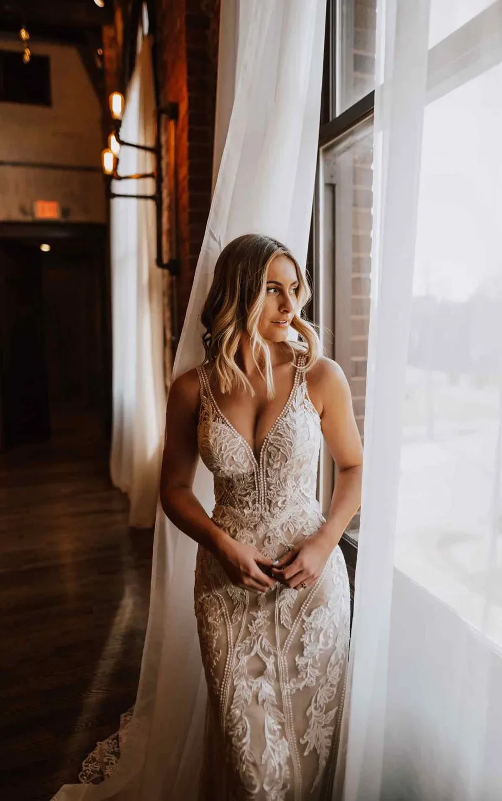 Braut steht in einem eleganten Hochzeitskleid mit tiefem V-Ausschnitt und Spitze vor einem Fenster und blickt nach draußen.