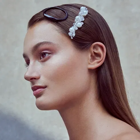 Seitliches Porträt einer Frau mit glattem, braunem Haar, die einen eleganten, perlenbesetzten Haarschmuck trägt. Ihr Make-up ist dezent und natürlich.